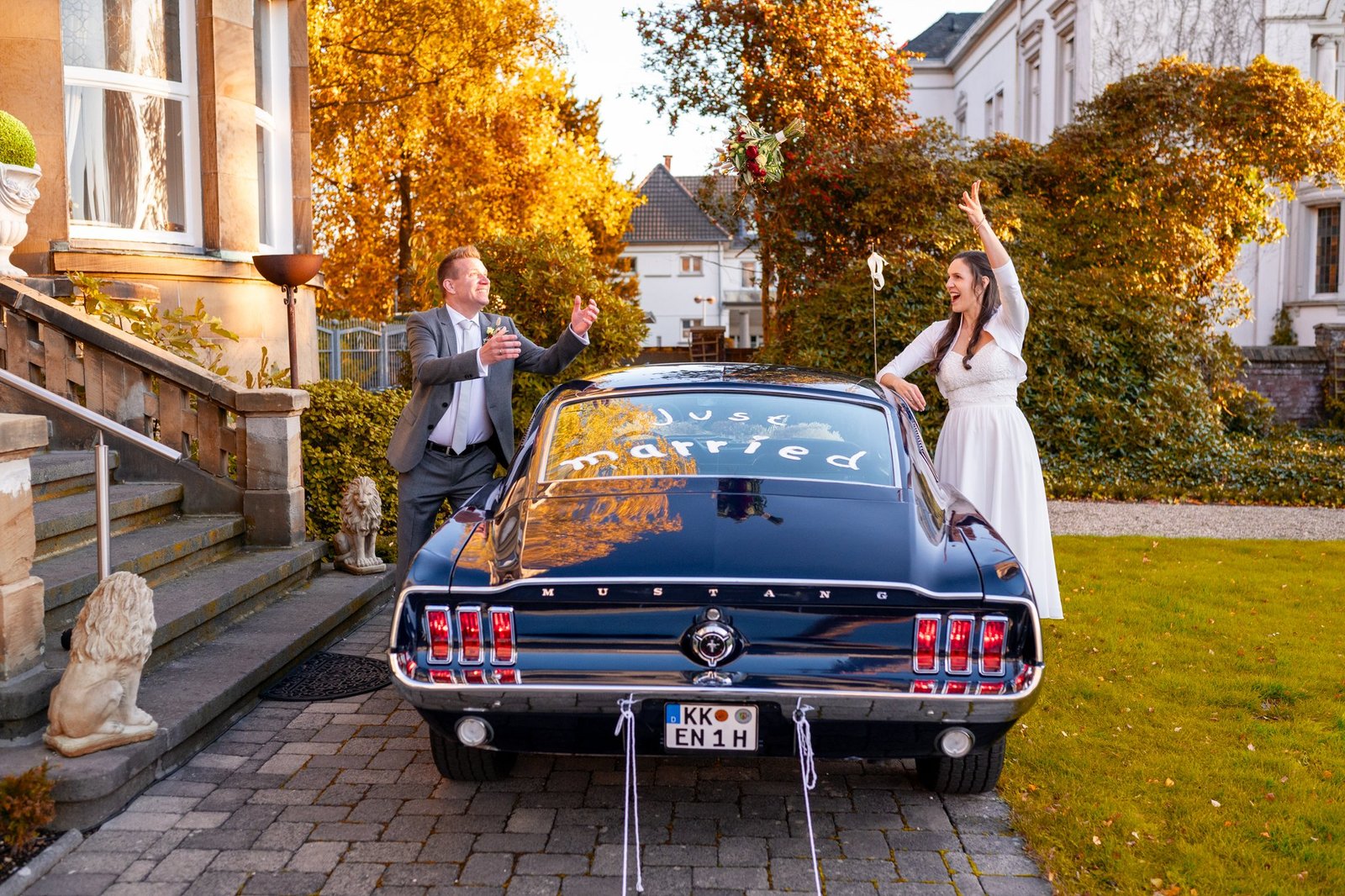 Laura und Enno werfen lachend den Brautstrauß über ihrem klassischen Mustang in die Luft